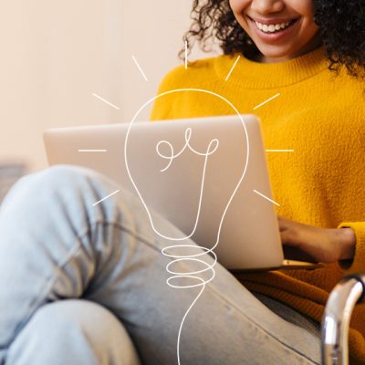 Uma mulher de camiseta amarela e cabelo cacheado, sentada de perna cruzada usando um notebook e sorrindo. Por cima da imagem, um desenho de lâmpada.